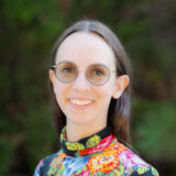 A person in a colorful floral shirt and tinted glasses poses for a photo in front of a wall of green foliage