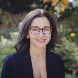 A person with red glasses and a navy blazer poses for a photo in front of a landscape of fall foliage and green leaves