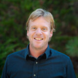 A person in a navy blue button-up shirt poses for a photo in front of a wall of green foliage