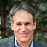 A person with short dark hair, wearing a gray suit, poses for a photo outdoors