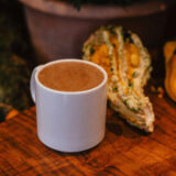 A white mug of hot chocolate sits on a wooden table next to an orange and yellow gourd