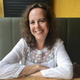 A person with long brown hair, wearing a white long-sleeve shirt, poses for a photo while sitting in a restaurant booth