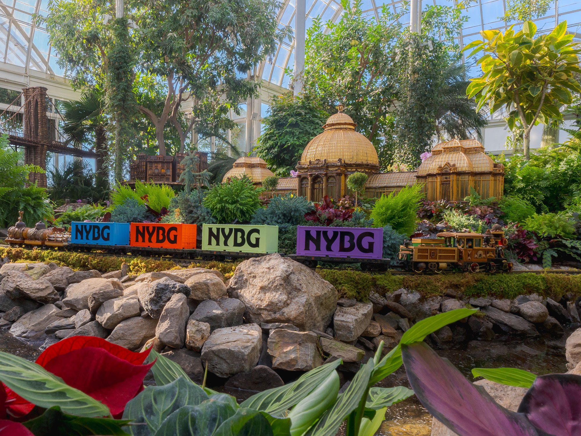 A conservatory setting featuring miniature buildings made of plant parts, with model trains zipping past