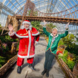 A person waves to the camera in a red Santa Claus suit with white curly hair and a hat. Next to him is a person in a green velvet jacket dressed as an elf with both hands extended above their head.