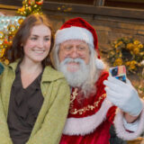 A person with long dark hair, wearing a green sweater and black shirt, poses for a photo with Santa Claus