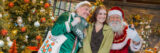 A person dressed as an elf in green, and on the other side a person with a long white beard dressed as Santa Claus in red and white, take a photo with a person wearing a green sweater in a festively decorated shop setting