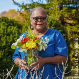 A person in a blue shirt poses for a photo in a sunny outdoor space, holding a vase of colorful flowers