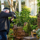 A person in a black jacket and hat uses a DSLR camera to photograph a miniature building display