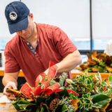 A person in a red shirt and blue hat puts together a wreathe of green leaves