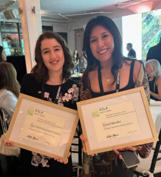 Two people in black dresses pose for a photo while holding framed award documents