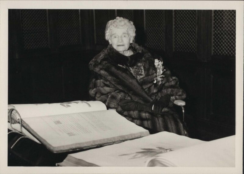 A black and white photo of a person in a dark fur coat photographed alongside two large, open books