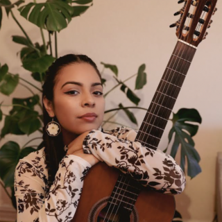 A person in a black and white floral shirt poses for a photo while holding an acoustic guitar