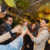 A group of people in a warmly lit conservatory after dark toast each other with cocktails