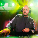 A person wearing headphones and a black tracksuit works DJ equipment under green stage lights