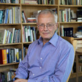 A person in a blue button-up shirt poses for a photo in front of a bookcase full of books