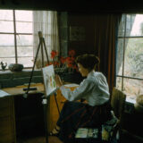 A person sits at an easel inside a home, painting