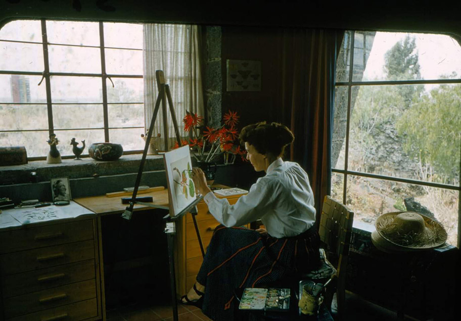 A person sits between two picture windows in a home, painting on an easel