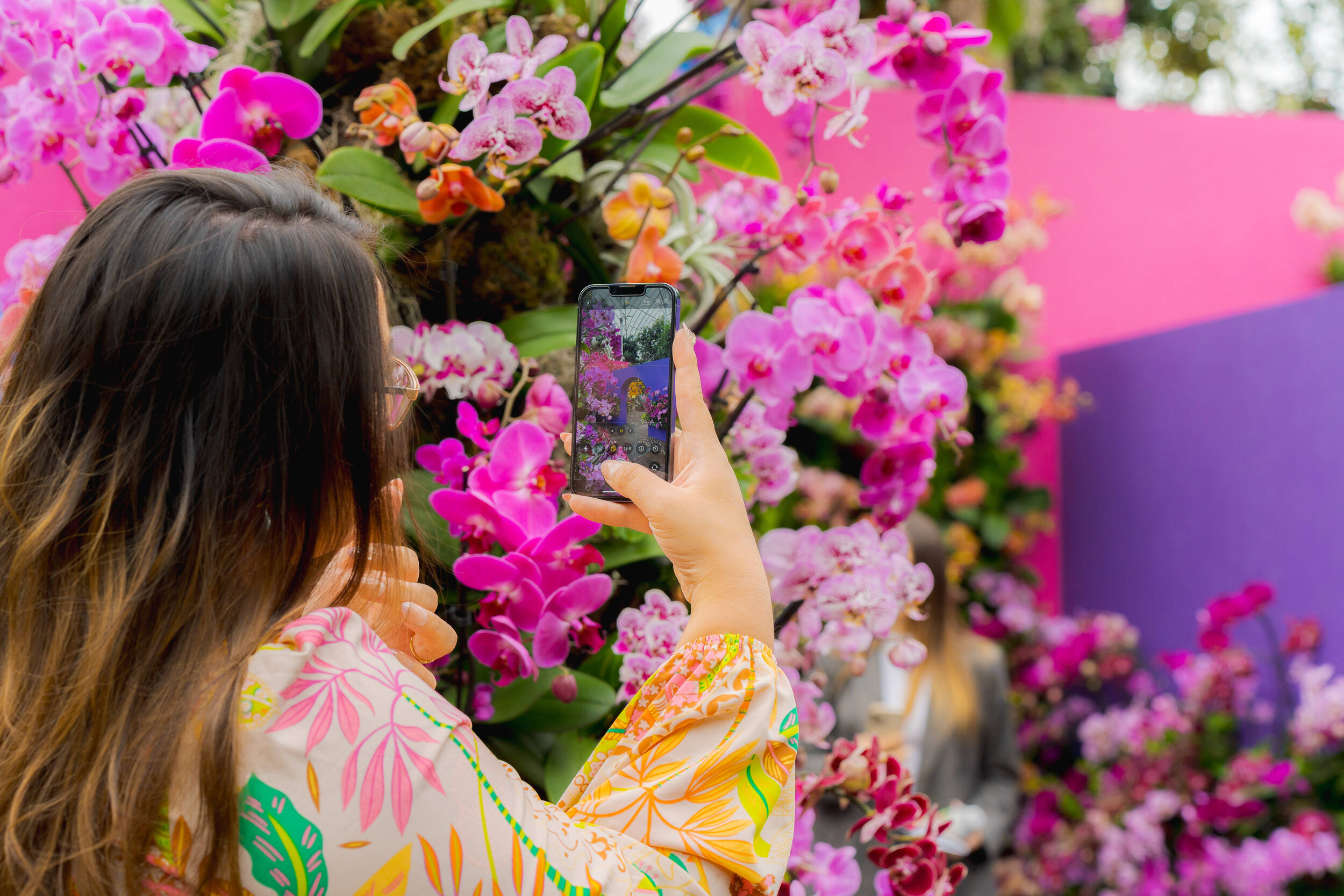 A person in a floral shirt, with long brown hair, takes a selfie among a profusion of pink and purple flowers