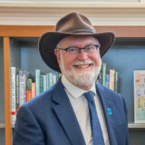 A person in a navy suit and brown brimmed hat poses for a photo in a library