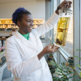 A person in a white lab coat examines a jar of preserved plant matter